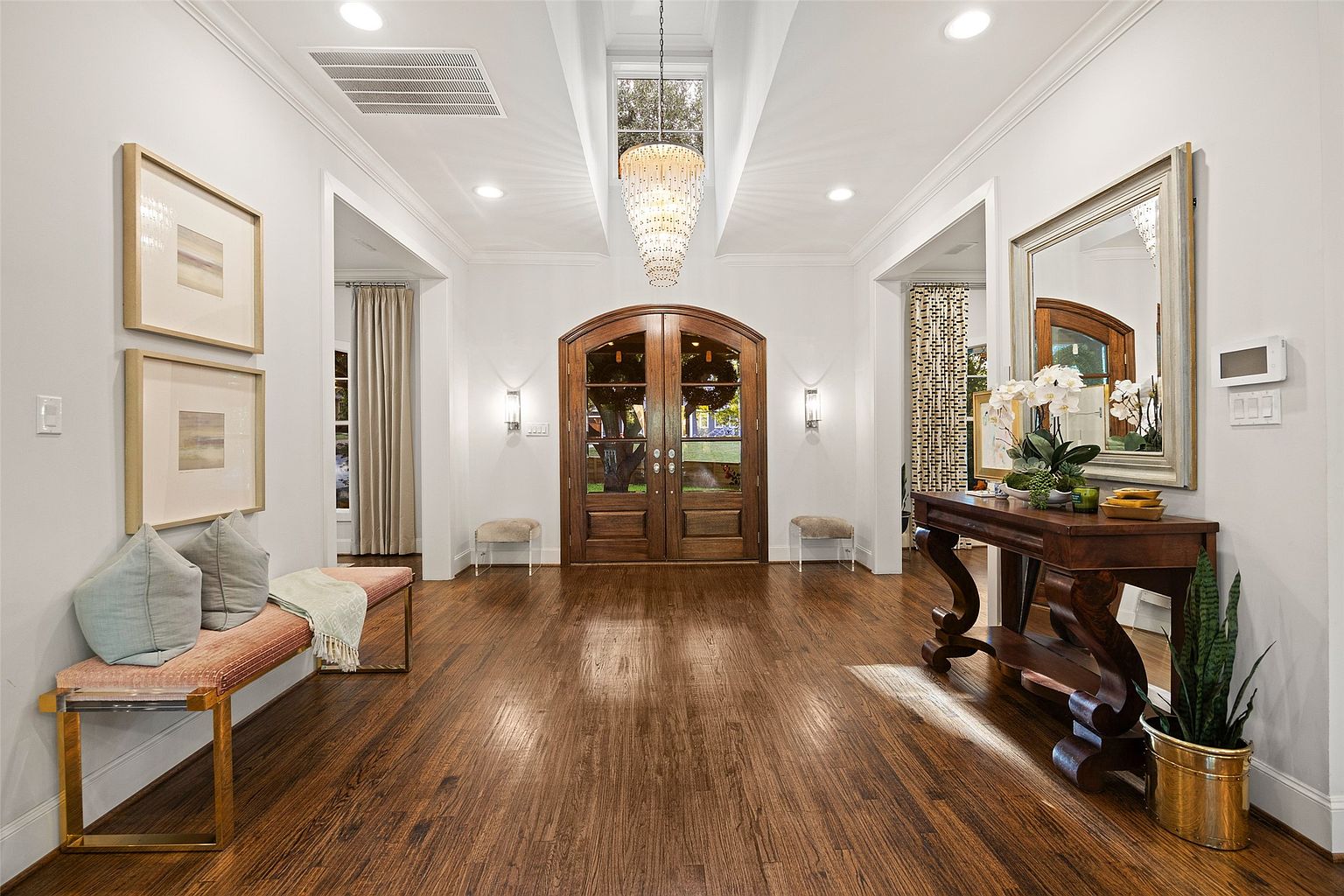 This is an interior shot of a grand entryway featuring dark hardwood floors, white walls, and a high ceiling with a decorative chandelier. A wooden console table with a mirror and floral arrangement sits to the right, while a bench with pillows is on the left. The perspective is from the center of the hallway, emphasizing the depth and spaciousness of the area.