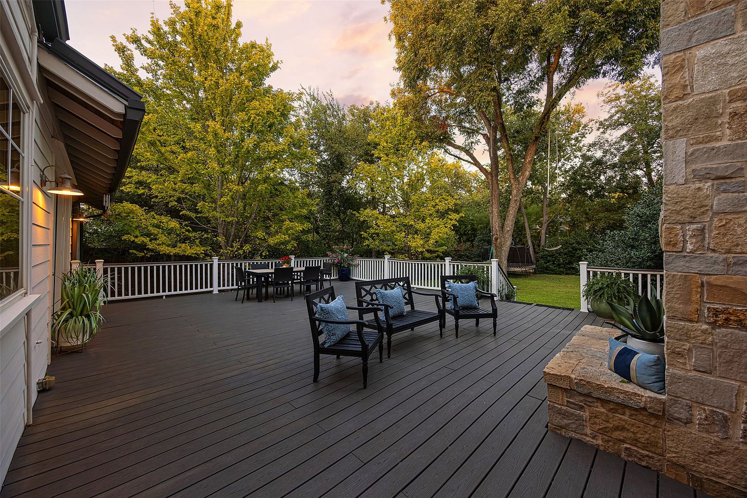 This image showcases a spacious and well-maintained deck area, perfect for outdoor entertaining. The deck features dark-stained wood planks, white railings, and comfortable seating arrangements with blue pillows. Lush greenery and mature trees surround the deck, creating a private and serene atmosphere. A stone structure with plants adds a touch of elegance and architectural interest.