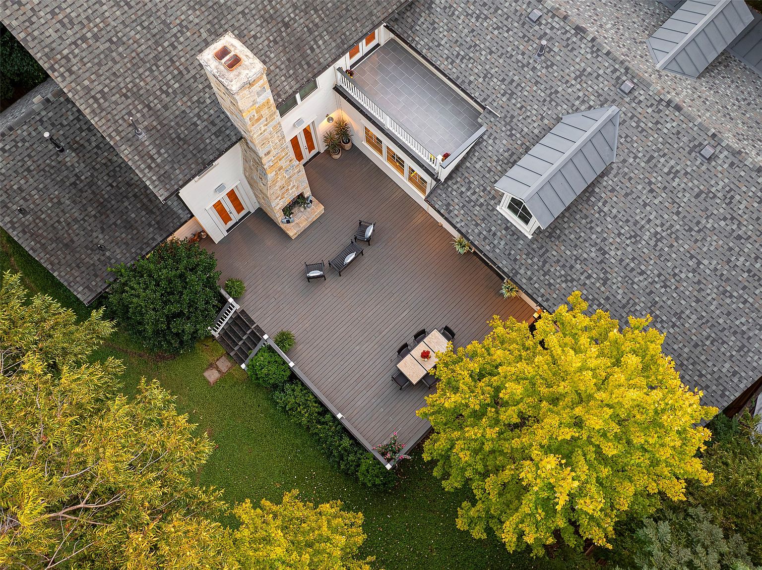 This aerial shot showcases the rear of a home with a spacious deck area featuring outdoor seating and dining. The deck is surrounded by lush greenery and mature trees, providing privacy and a serene atmosphere. The roof is a mix of gray shingles with a unique dormer window, and a stone chimney adds a touch of rustic charm.