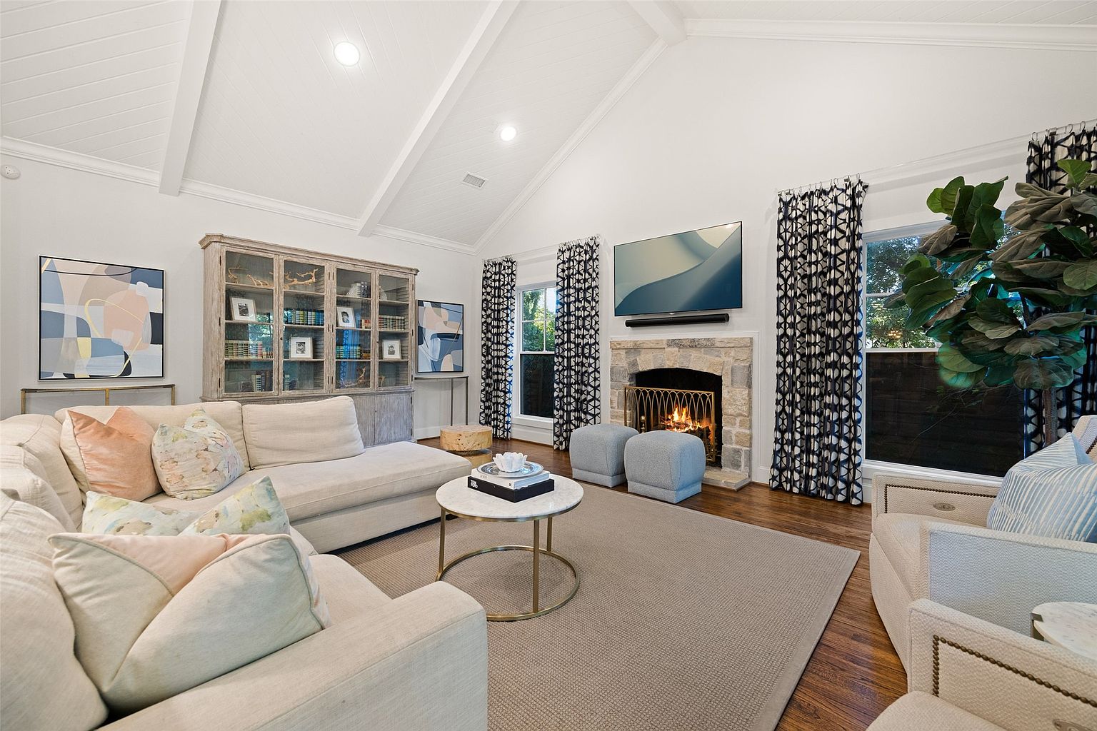 This is a bright and airy living room featuring a vaulted ceiling with exposed beams, a stone fireplace with a mounted television, and large windows with patterned curtains. The room is furnished with a comfortable sectional sofa, a round coffee table, and two upholstered armchairs, creating a cozy and inviting atmosphere. The hardwood floors and neutral-toned rug add warmth and texture to the space.