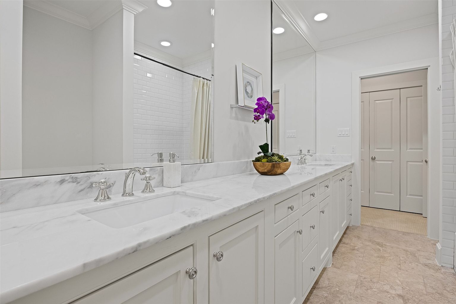 This is a bright and elegant primary bathroom featuring a long vanity with a white marble countertop and white cabinetry. The large mirror reflects the shower area and enhances the sense of space. The room is well-lit with recessed lighting and showcases a clean, modern design with classic touches.