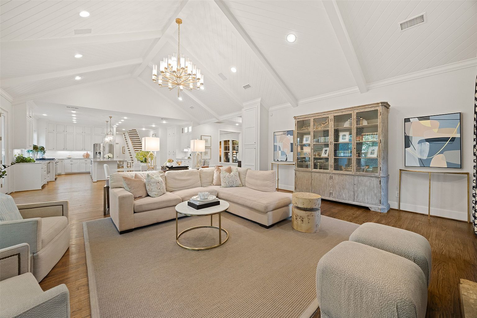 This is a bright and airy living room featuring a large sectional sofa, a round coffee table, and a neutral-toned rug. The room is well-lit by a chandelier and recessed lighting, highlighting the vaulted ceiling and hardwood floors. A large cabinet and artwork add character to the space, creating a comfortable and inviting atmosphere.