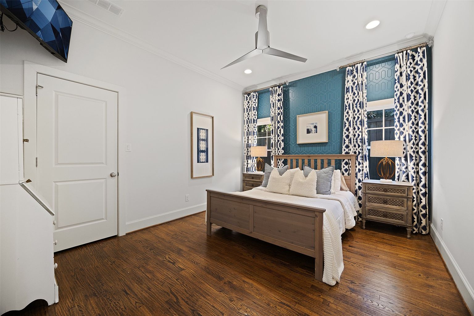 This is a well-lit primary bedroom featuring a wooden bed frame with neutral-toned pillows and a white throw blanket. The room has hardwood floors, white walls, and a blue patterned accent wall behind the bed, flanked by windows with matching curtains. Two nightstands with lamps sit on either side of the bed, and artwork is displayed on the walls, creating a cozy and inviting atmosphere.