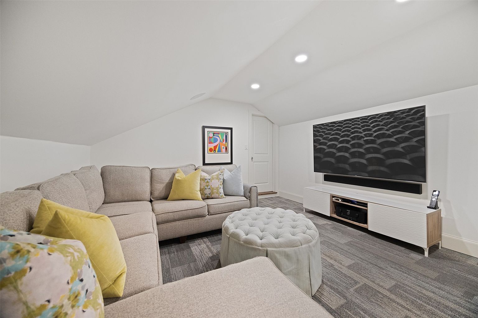 This is a cozy living room featuring a sectional sofa with decorative pillows, a tufted ottoman, and a large flat-screen TV mounted above a modern media console. The room has a neutral color palette with gray carpet and white walls, creating a clean and inviting atmosphere. A colorful framed artwork adds a touch of personality to the space.