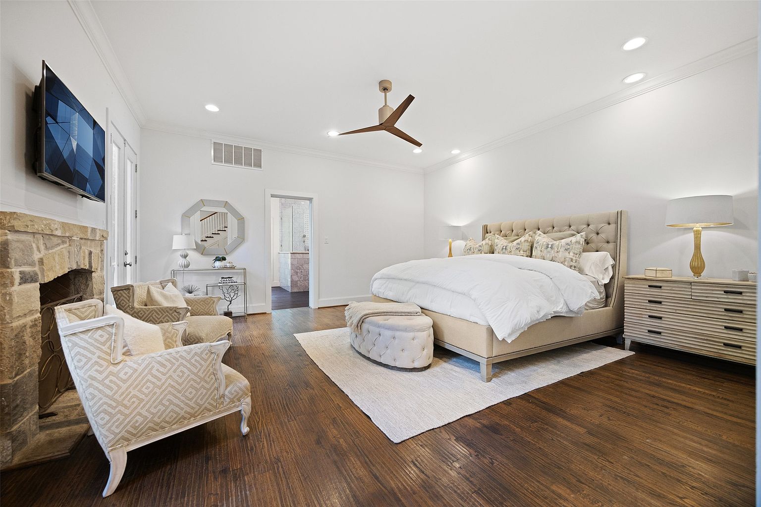 This is a primary bedroom featuring a large bed with a tufted headboard, a neutral color palette, and hardwood floors. The room includes a fireplace with a TV mounted above, a seating area with patterned armchairs, and a dresser with a lamp. The overall impression is luxurious and comfortable.