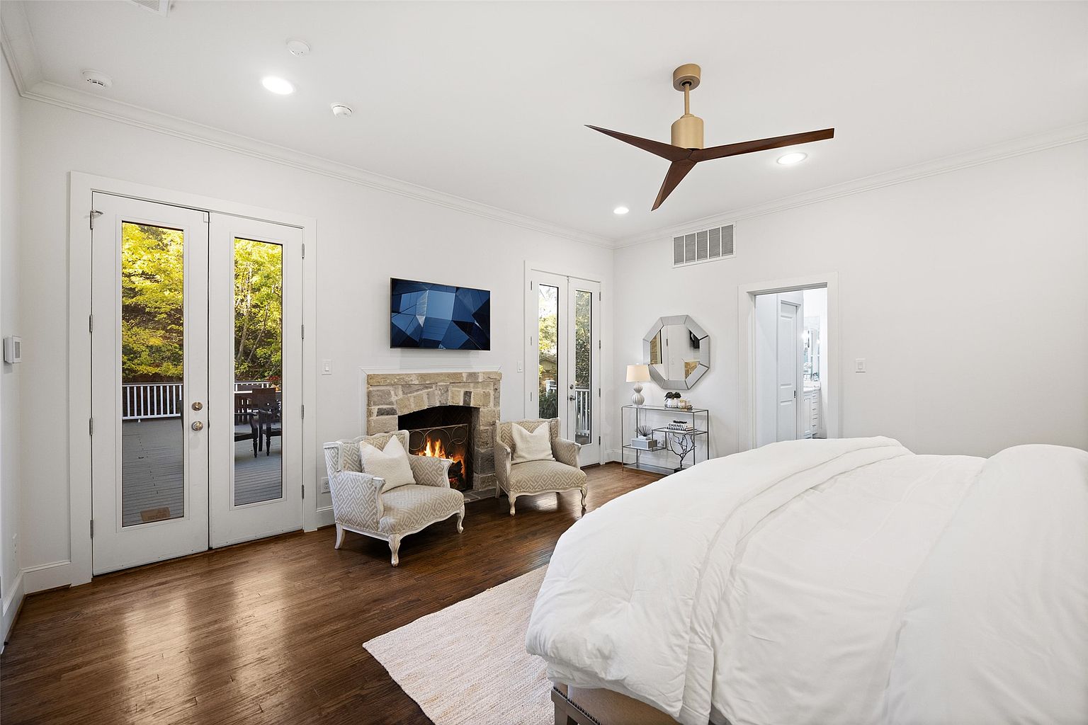 This is a bright and airy primary bedroom featuring hardwood floors, white walls, and a stone fireplace with a mounted television above. The room includes two upholstered armchairs, a ceiling fan, and French doors leading to an outdoor deck. A large bed with white linens dominates the foreground, creating a cozy and inviting atmosphere.