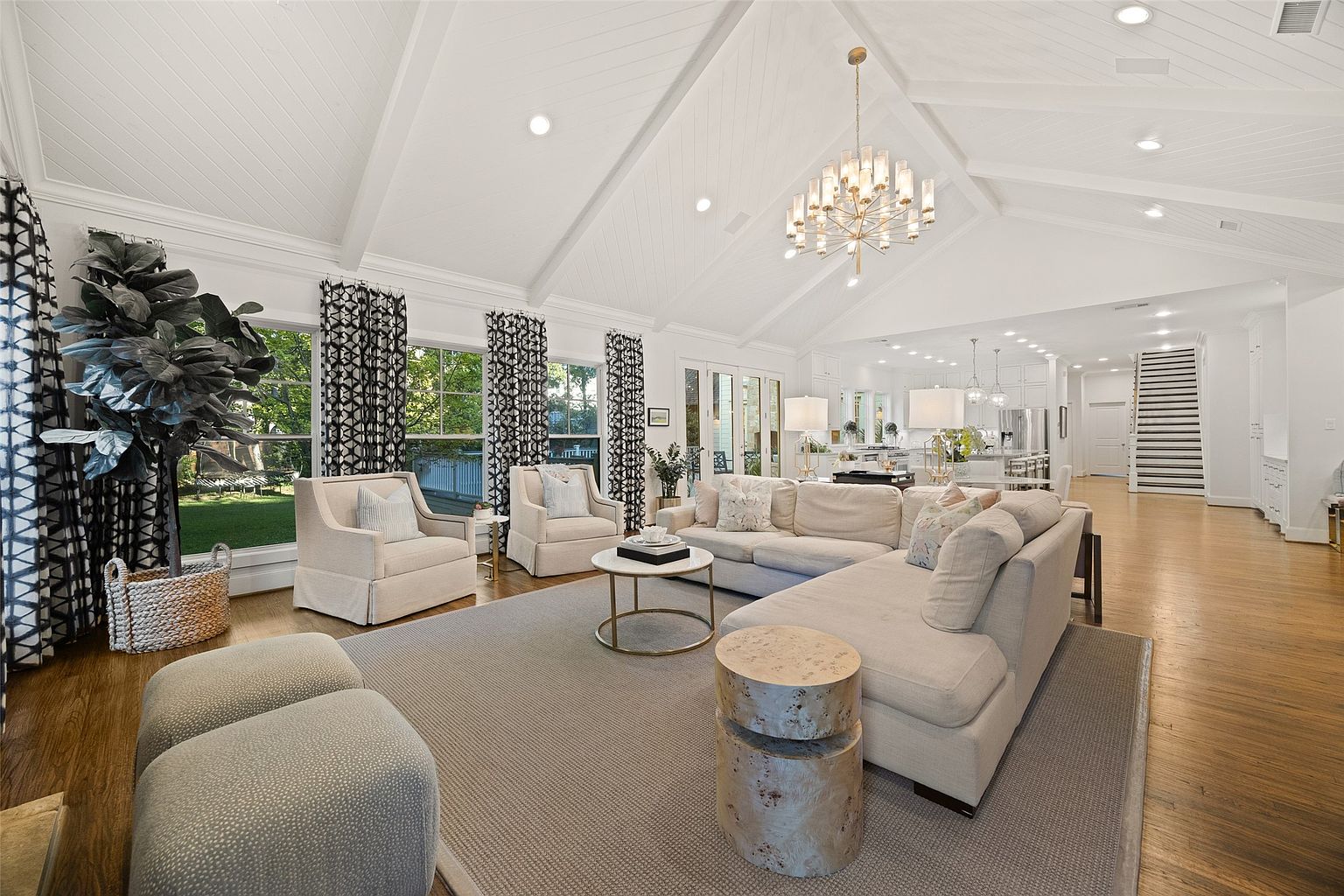This is a bright and spacious living room featuring a high vaulted ceiling with exposed beams and recessed lighting. The room is furnished with a large sectional sofa, two armchairs, and a round coffee table, all arranged on a large area rug. Natural light floods the space through large windows dressed with patterned curtains, creating a welcoming and airy atmosphere.