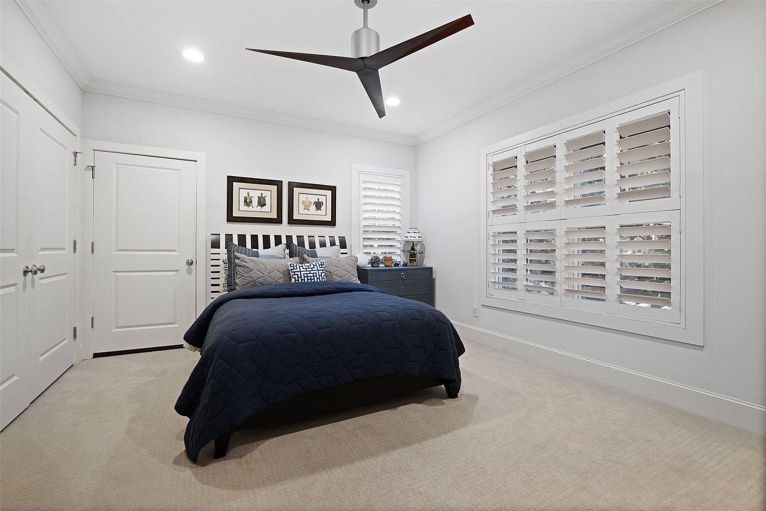 This is a bright and airy bedroom featuring a bed with a navy blue geometric patterned quilt, a striped headboard, and decorative pillows. The room has white walls, white trim, and a light beige carpet. Natural light streams in through the window with white shutters, and a modern ceiling fan adds a stylish touch. Two framed pictures hang above the bed.