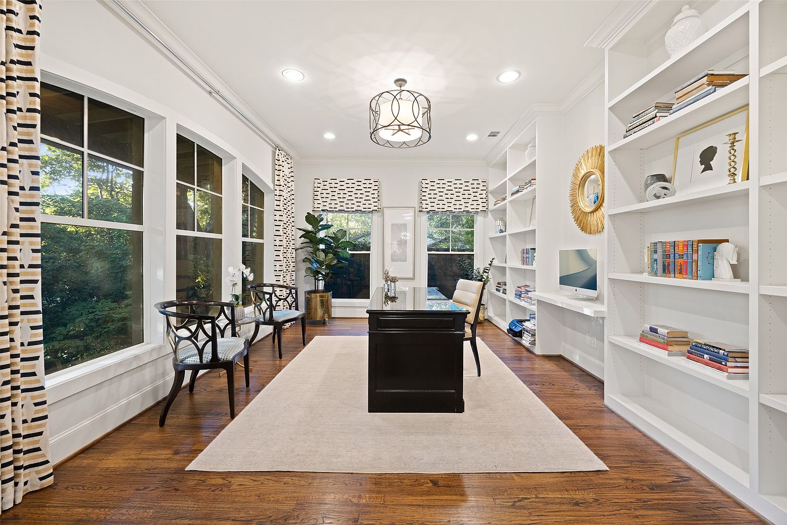 This is an interior shot of a well-lit home office. The room features hardwood floors, a large area rug, and built-in bookshelves. A dark wood desk sits in the center of the room, and large windows provide natural light. The overall impression is one of sophistication and functionality.