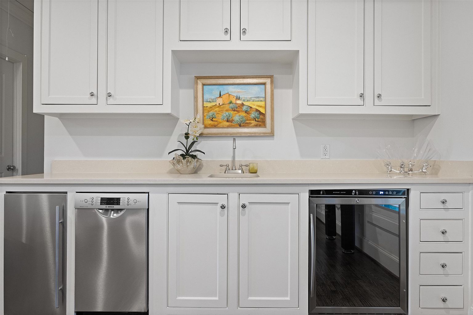 This is a well-lit kitchen featuring white cabinetry with silver hardware, stainless steel appliances including a dishwasher and beverage cooler, and light-colored countertops. A painting hangs above the sink, adding a touch of color to the space. The perspective is a medium shot, showcasing the functionality and design of the kitchen area.