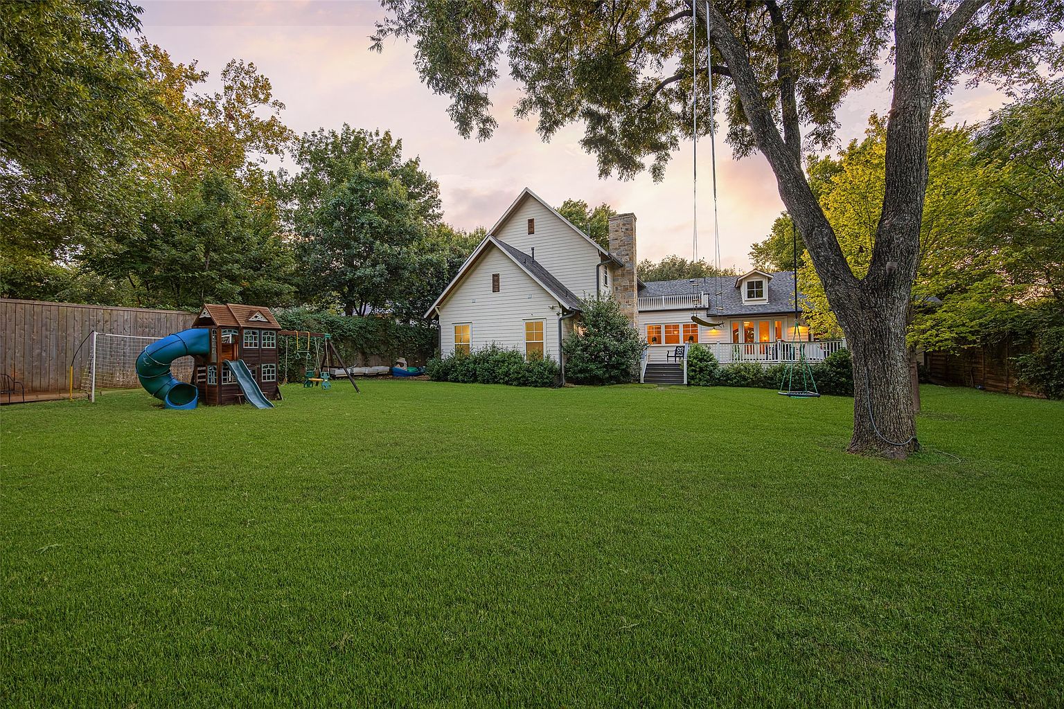 This image showcases a spacious backyard with a well-maintained lawn, a playset with a slide, and mature trees providing shade. The house is visible in the background, featuring a deck and a chimney. The overall impression is a family-friendly and inviting outdoor space.
