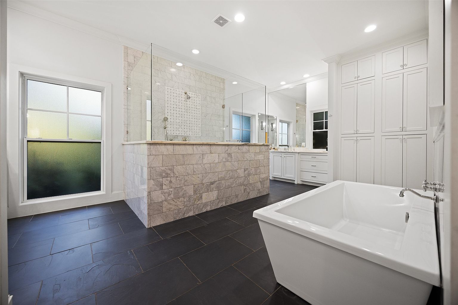 This is a bright and spacious primary bathroom featuring dark tile flooring and white walls. A large, modern bathtub sits in the foreground, while a walk-in shower with glass enclosure and marble-like tiling is visible in the background. White cabinetry provides ample storage, and the overall design exudes a clean and luxurious feel.