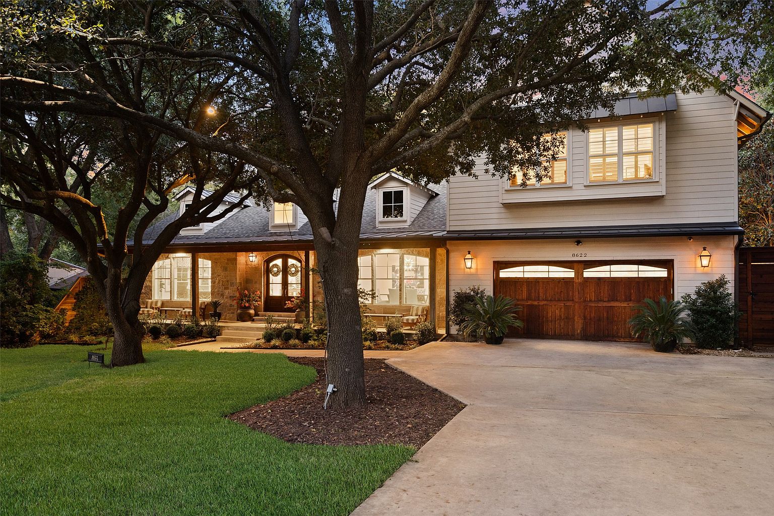 This is a front exterior view of a two-story house with a well-manicured lawn and mature trees. The house features a combination of stone and siding, a covered front porch, and a two-car garage with wooden doors. Exterior lighting fixtures add a warm glow, enhancing the curb appeal.