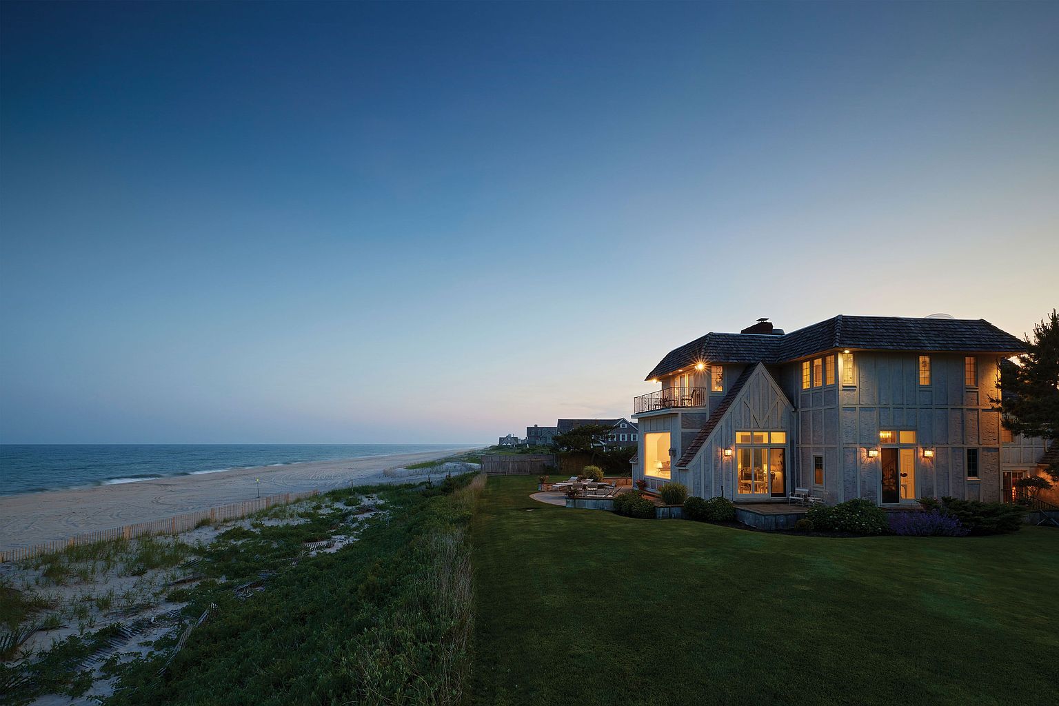 This image showcases the rear exterior of a luxurious beachfront home at dusk. The house features a multi-level design with a balcony, large windows, and a well-manicured lawn that extends to the sandy beach. The overall impression is one of coastal elegance and serene living.