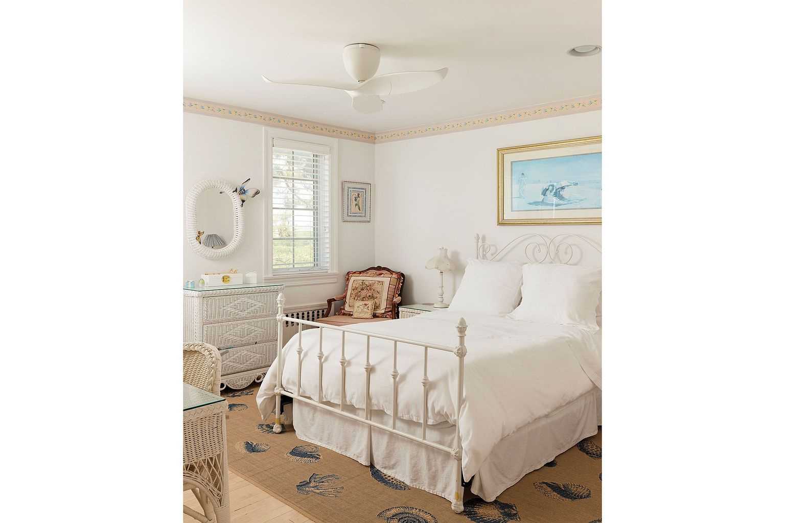This is a charming bedroom featuring a white iron bed with white linens, a white wicker dresser with a mirror, and a patterned rug. The room has a light and airy feel, enhanced by natural light from the window and a ceiling fan. The overall style is coastal and inviting, perfect for a guest room.
