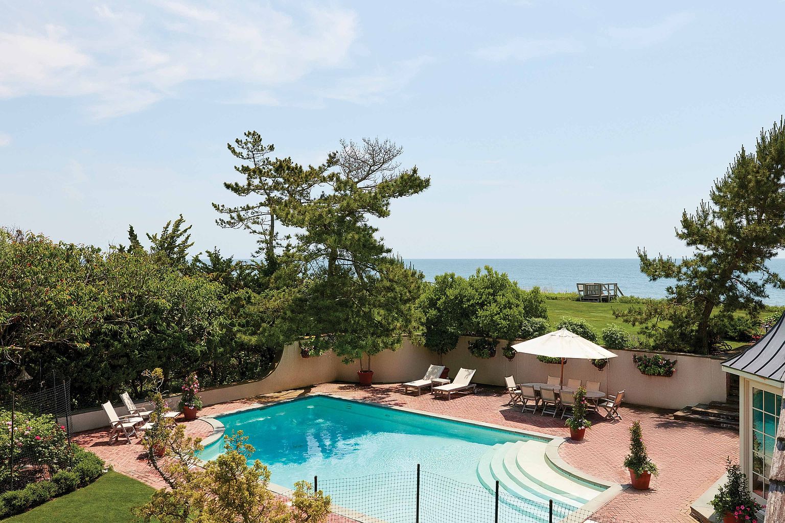 This image showcases a luxurious backyard pool area, featuring a rectangular pool surrounded by a brick patio. Lounge chairs and a dining set with an umbrella offer ample space for relaxation and entertainment. The area is enclosed by a low wall and lush greenery, providing privacy and a serene atmosphere, with a glimpse of the ocean in the background.
