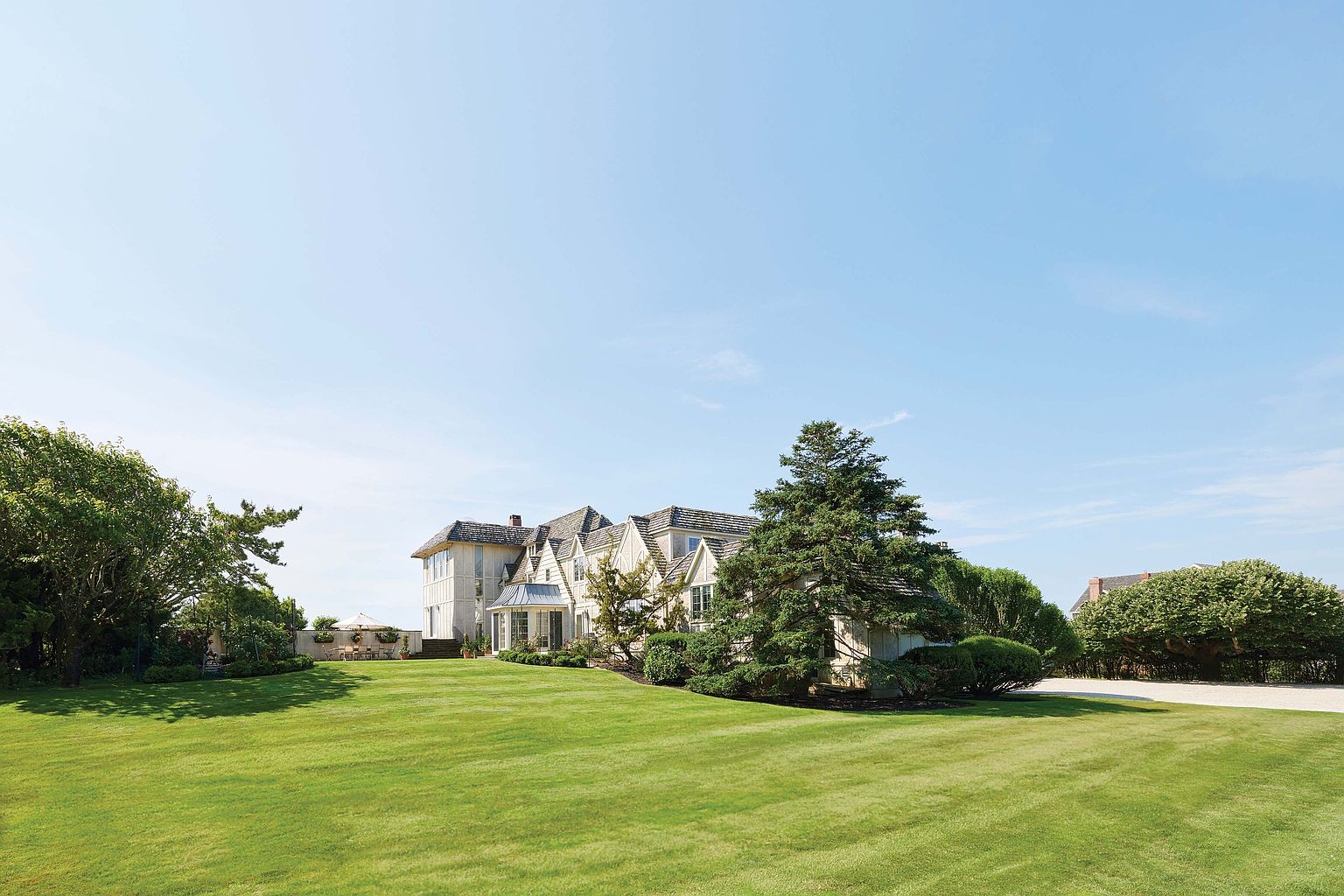 This image showcases a sprawling, meticulously maintained lawn leading up to a grand residence. The house features a classic architectural style with multiple gables and a light-colored exterior, complemented by mature trees and landscaping. The overall impression is one of luxury and spaciousness, highlighting the property's extensive outdoor space.