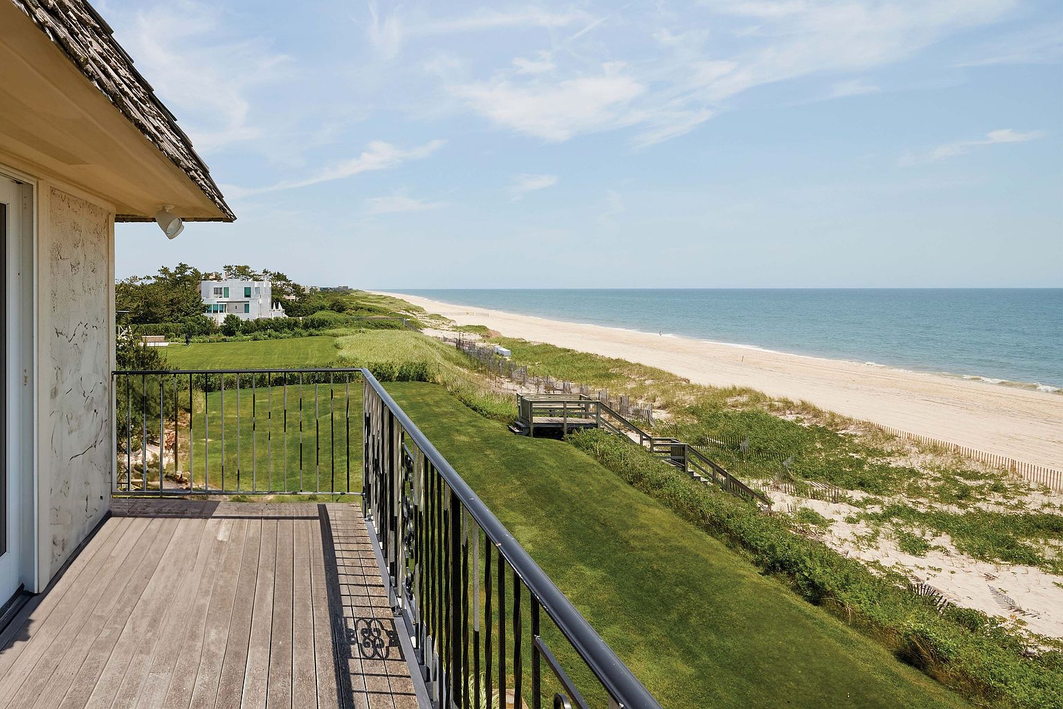 This image showcases a beautiful balcony overlooking a lush green lawn that extends to a sandy beach and the ocean. The balcony features a wooden deck and a black metal railing. In the distance, a modern white house is visible, adding to the property's appeal and highlighting its proximity to the beach.