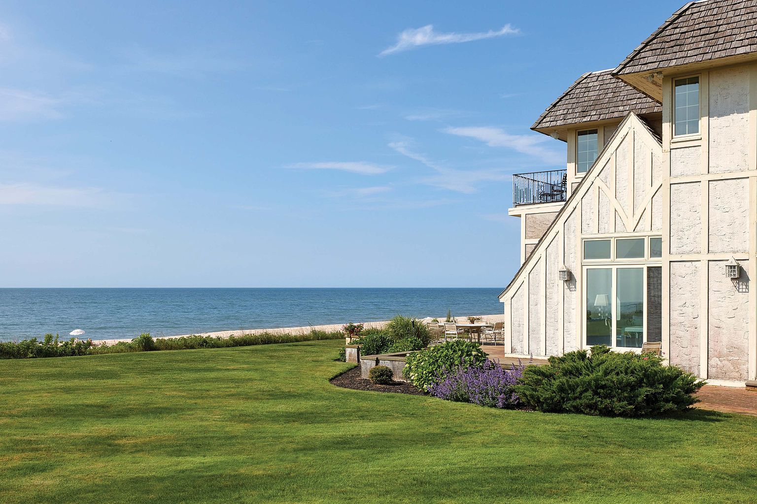 This image showcases a beautifully landscaped yard with a well-maintained lawn, vibrant flower beds, and mature shrubs, leading up to a stately home. The property overlooks a serene beach and ocean, creating a tranquil and luxurious setting. A patio area with outdoor furniture suggests an ideal space for relaxation and entertaining.