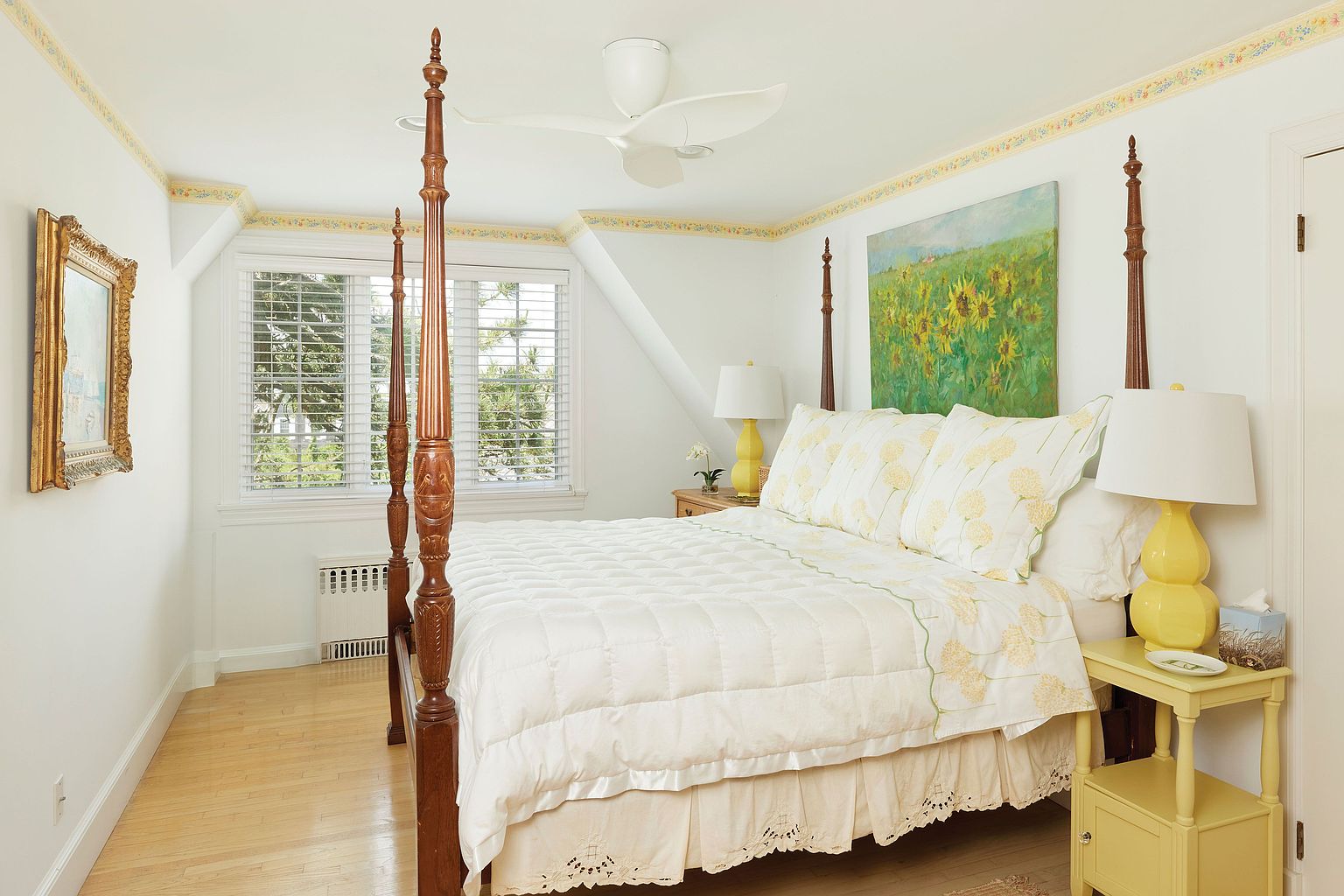 This is a bright and inviting primary bedroom featuring a four-poster bed with a white quilted comforter and floral patterned pillows. The room is decorated in a classic style with yellow accents, including bedside lamps and a small side table. Natural light streams in through the window, creating a warm and welcoming atmosphere.