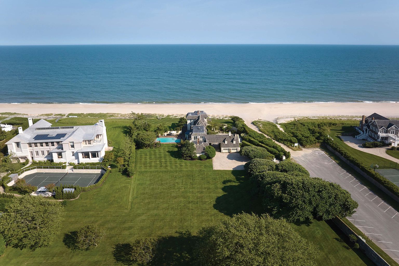 This aerial shot showcases a luxurious beachfront property featuring multiple houses with manicured lawns, a swimming pool, and a tennis court. The houses exhibit classic architectural styles, and the expansive green lawn extends towards the sandy beach and the ocean. A parking lot is visible on the right side of the image.