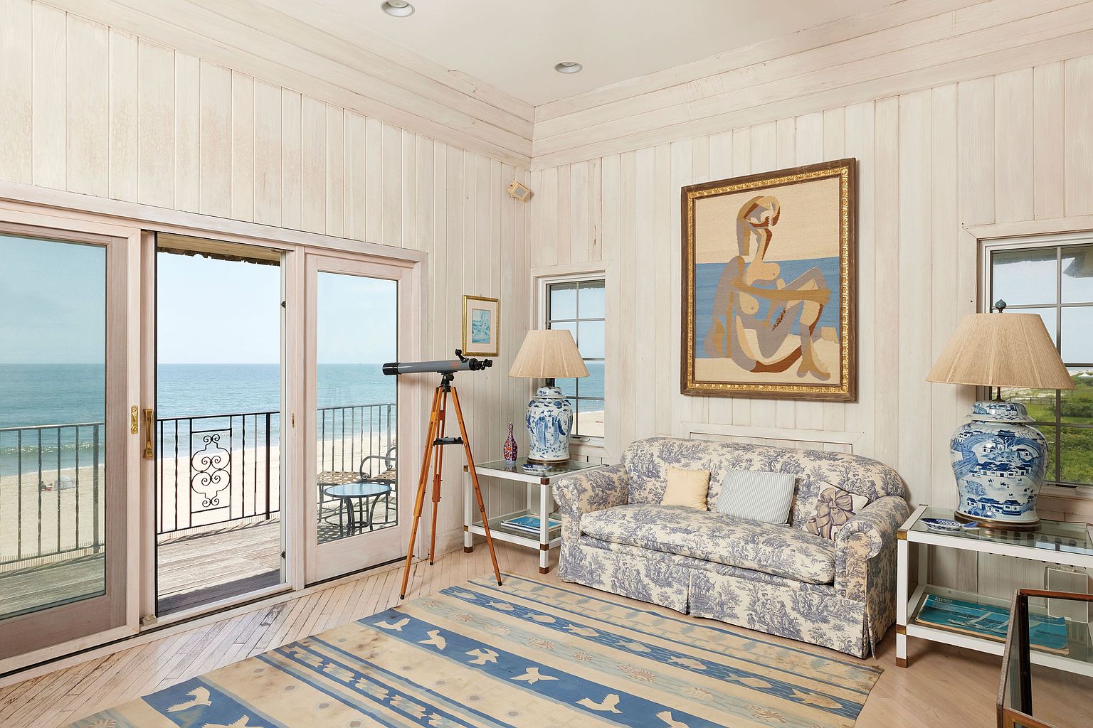This is an inviting living room with a coastal aesthetic, featuring light wood paneling and a blue and beige patterned rug. A comfortable sofa with matching upholstery sits beneath a framed artwork, flanked by glass-topped side tables with blue and white porcelain lamps. Sliding glass doors open to a balcony overlooking the ocean, enhancing the room's serene atmosphere.