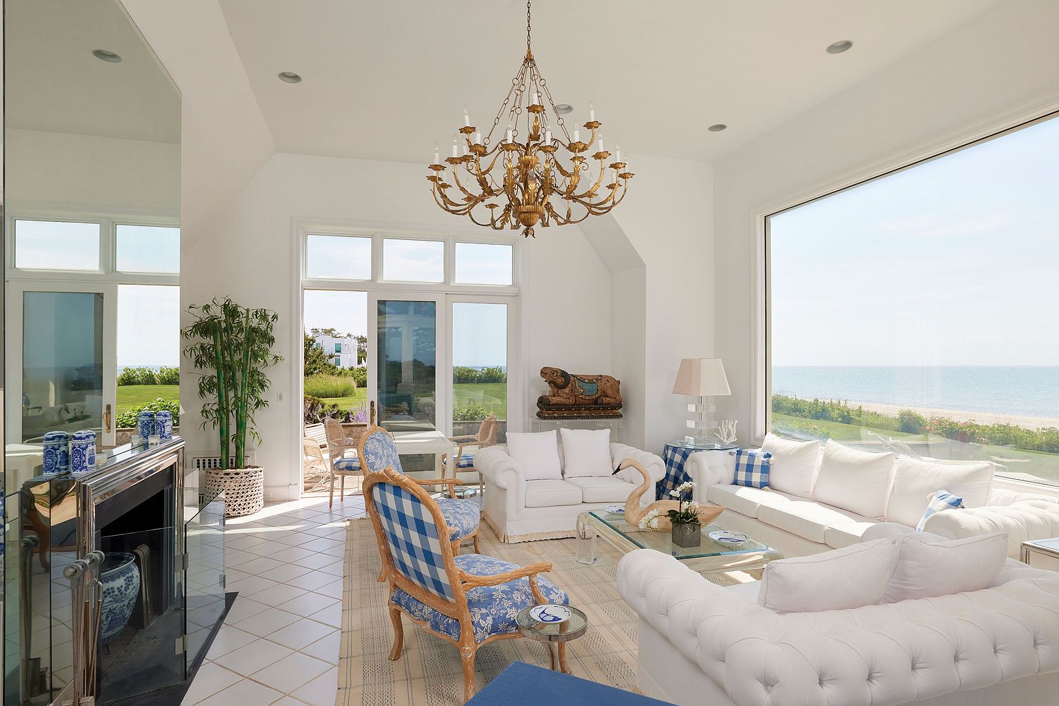 This is a bright and airy living room featuring white walls, tile flooring, and large windows offering ocean views. The room is furnished with white sofas, blue and white patterned chairs, and a gold chandelier, creating a luxurious yet comfortable atmosphere. A mirrored fireplace and potted plant add to the room's elegance.