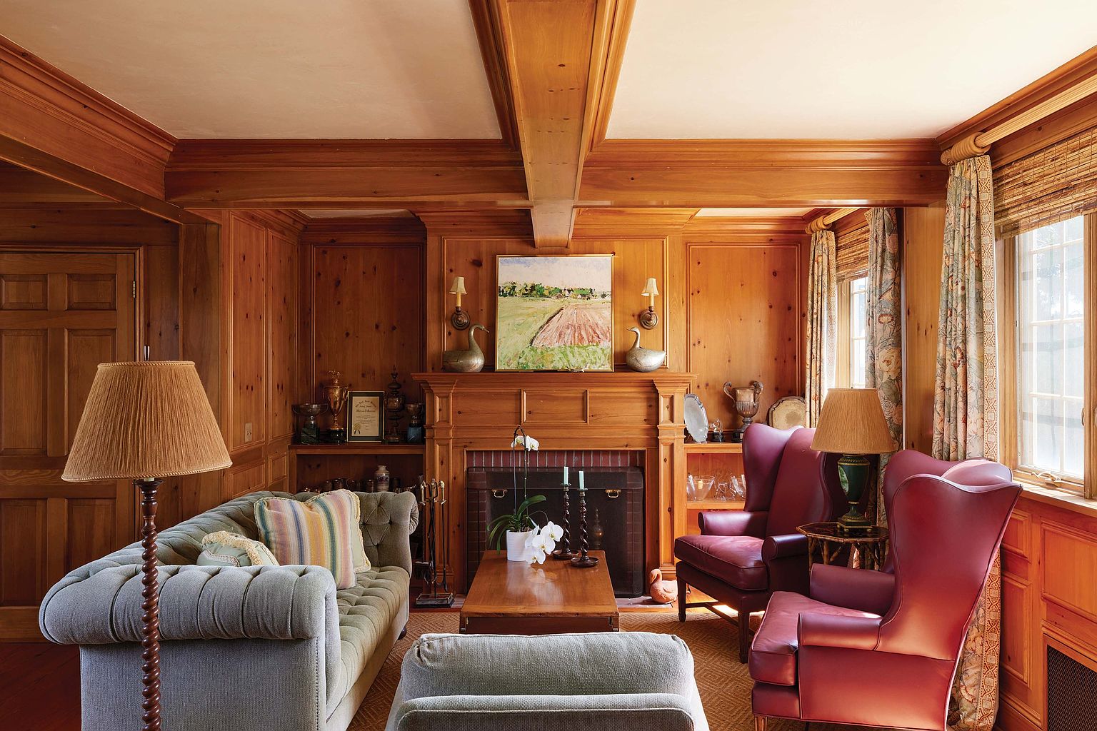 This is a warm and inviting living room featuring wood paneling and ceiling beams, creating a cozy and traditional atmosphere. The room is furnished with a tufted sofa, two red wingback chairs, and a wooden coffee table, complemented by lamps and decorative items. A fireplace with a mantel and artwork above it serves as a focal point, enhancing the room's charm and character.