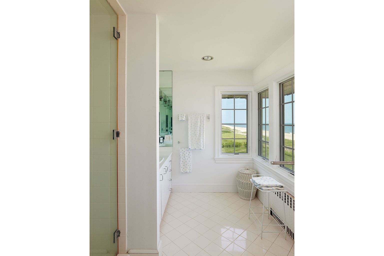 This is a bright and airy bathroom featuring a walk-in shower with glass doors, a white vanity with a mirror, and a set of windows offering a view of the ocean. The bathroom has a clean and simple design with white walls and tiled floors, creating a serene and relaxing atmosphere. The perspective is from the doorway, looking into the bathroom.
