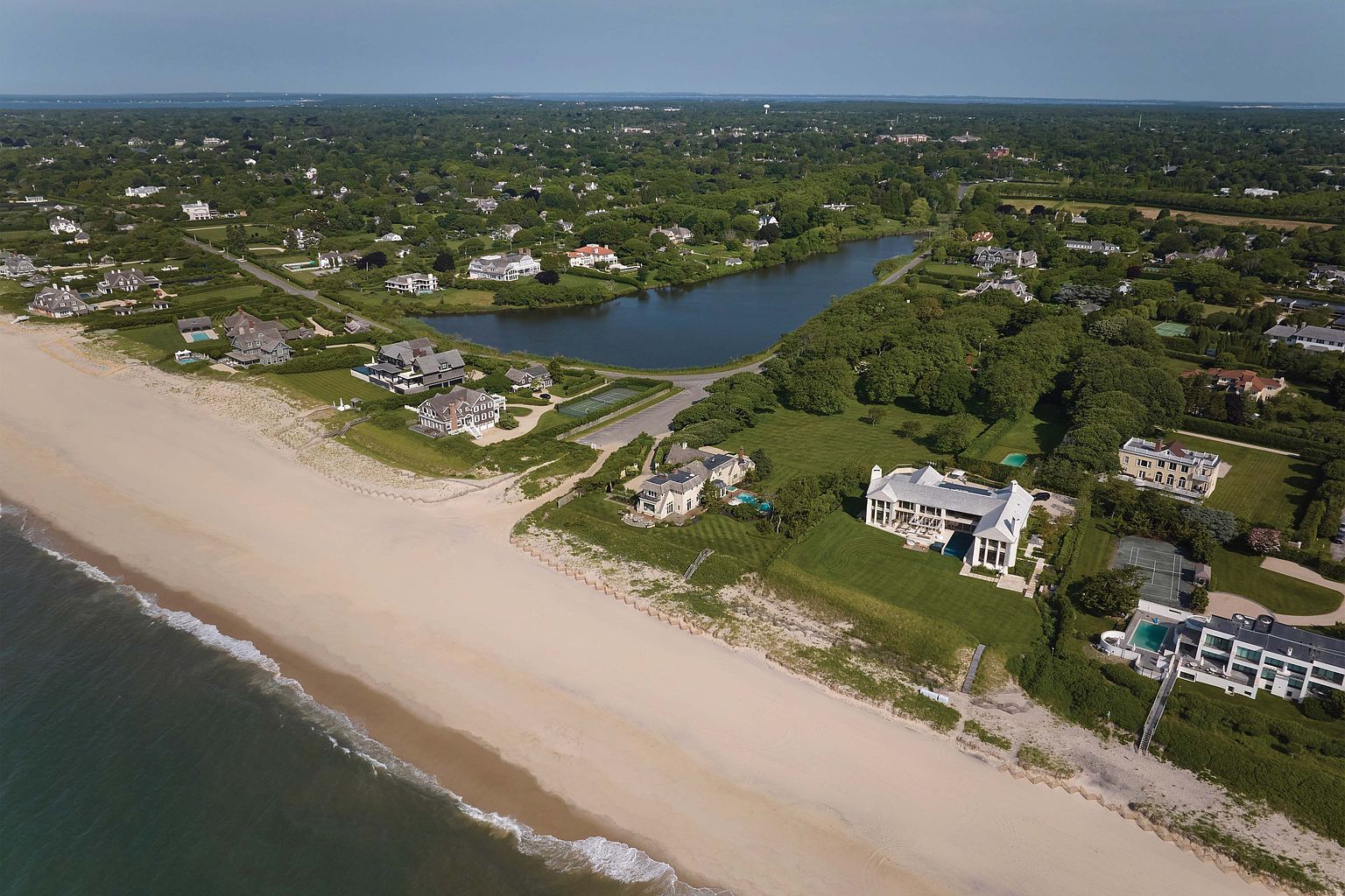 This aerial view showcases a luxurious coastal property featuring a grand white mansion with manicured lawns extending to a pristine sandy beach. The property includes a swimming pool, tennis court, and multiple structures, all nestled amidst lush greenery and overlooking a serene body of water. The perspective offers a cinematic overview of the estate's scale and prime location.