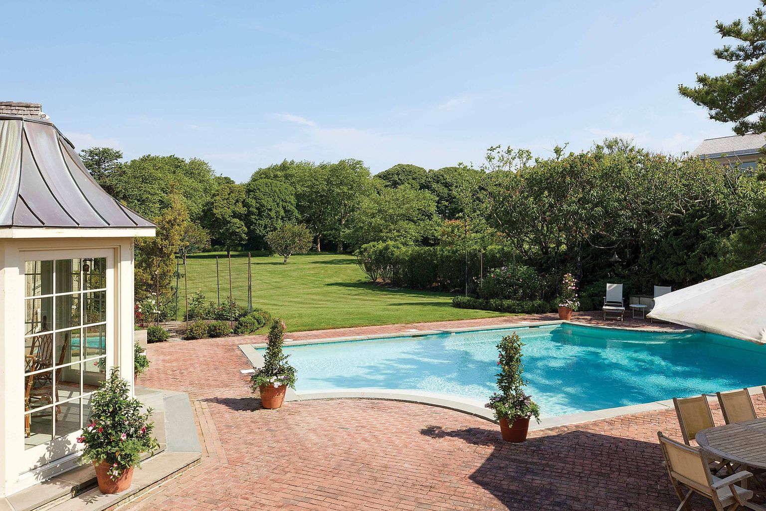 This image showcases a luxurious backyard pool area, featuring a pristine blue pool surrounded by a red brick patio. The pool is flanked by potted plants, and a well-manicured lawn extends into the distance, bordered by lush trees and greenery. A covered outdoor dining area is visible, suggesting an ideal space for relaxation and entertainment.