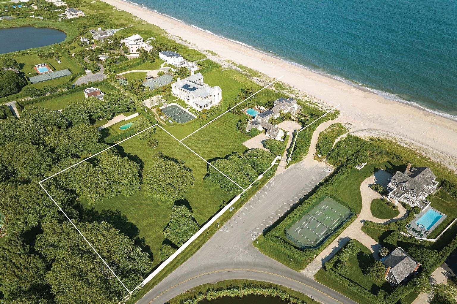 This aerial view showcases a luxurious beachfront property with meticulously manicured lawns, a tennis court, and a swimming pool. The expansive estate includes multiple buildings, mature trees, and direct access to a pristine sandy beach and the ocean. The perspective is from above, providing a comprehensive view of the property's layout and surrounding landscape.