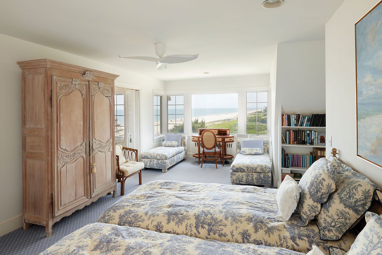 This primary bedroom features a serene coastal aesthetic with a large bed adorned in blue and white toile fabric, complemented by matching chaise lounges near a window offering ocean views. A light wood armoire adds a touch of rustic charm, while a built-in bookshelf and a painting contribute to the room's sophisticated ambiance. The room is bright and airy, creating a relaxing retreat.