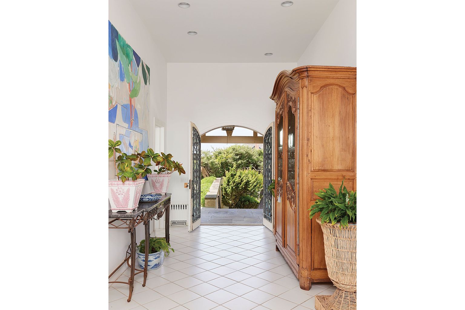This interior shot showcases a bright and airy hallway with white tiled floors and walls, leading to an open doorway with a view of a lush garden. The hallway is decorated with a large abstract painting, a console table with potted plants, and a large wooden cabinet, creating a welcoming and elegant entryway. The perspective is from the entrance of the hallway, drawing the viewer's eye towards the garden view.