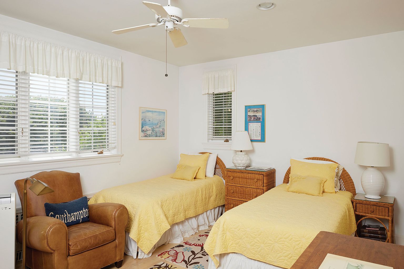 This is a bright and inviting guest bedroom featuring two twin beds with yellow quilted bedspreads and matching pillows. The room is painted in a clean white, complemented by white window blinds and curtains. A comfortable leather armchair sits in the corner, adding a cozy touch to the space.