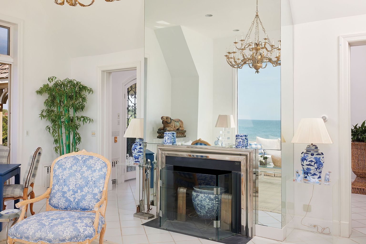 This interior shot showcases a bright and airy living room with a sophisticated design. The room features a mirrored wall reflecting an ocean view, a decorative fireplace, and elegant furnishings, including a blue floral armchair. The white tile flooring and neutral walls contribute to a clean and spacious feel, perfect for a luxurious coastal home.