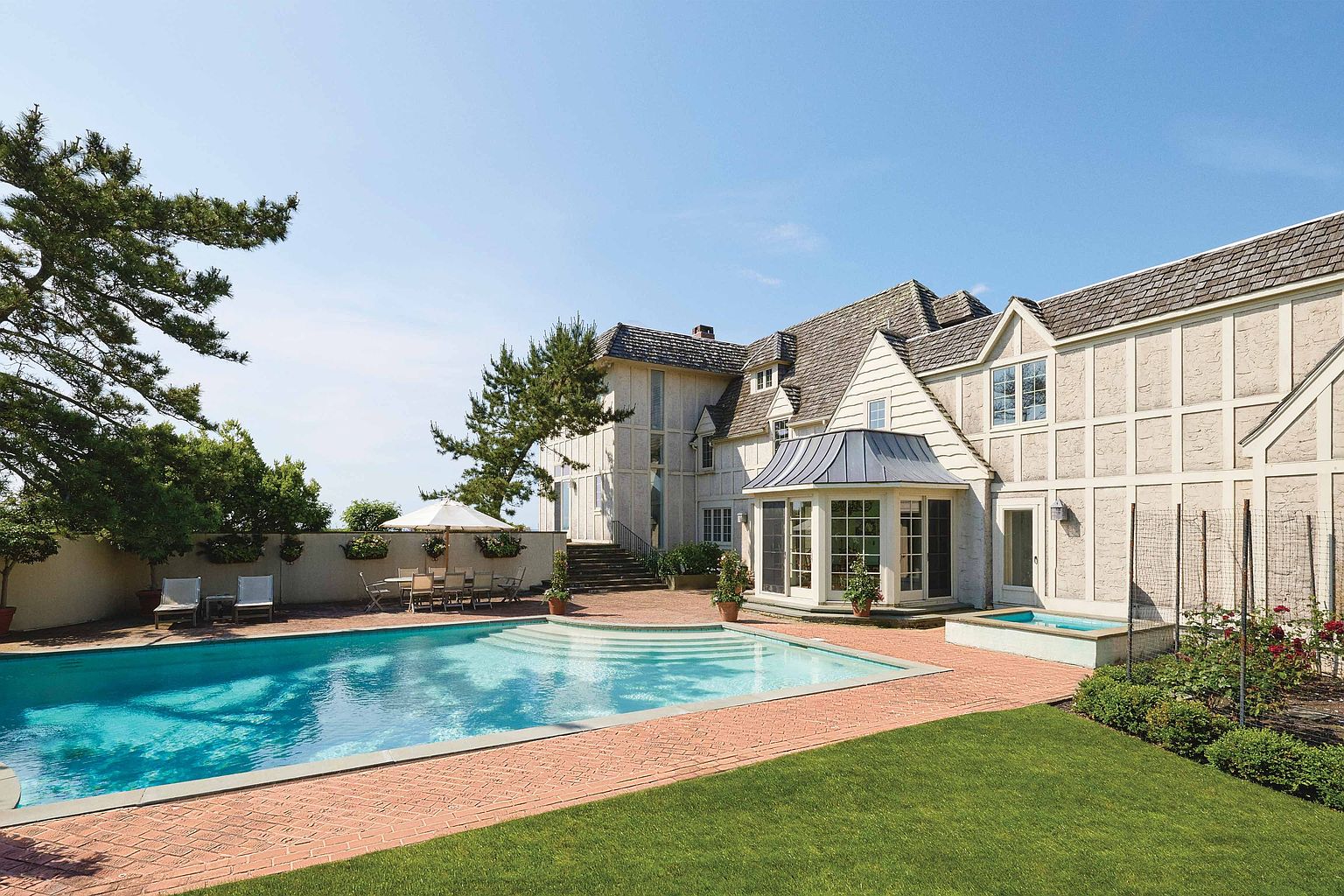 This image showcases the rear exterior of a luxurious home, emphasizing the inviting pool and spa area. The pool features a gradual entry with steps, surrounded by a brick patio and lush green lawn. The house itself exhibits a classic architectural style with a combination of stucco and timber detailing, complemented by a sunroom with a metal roof.