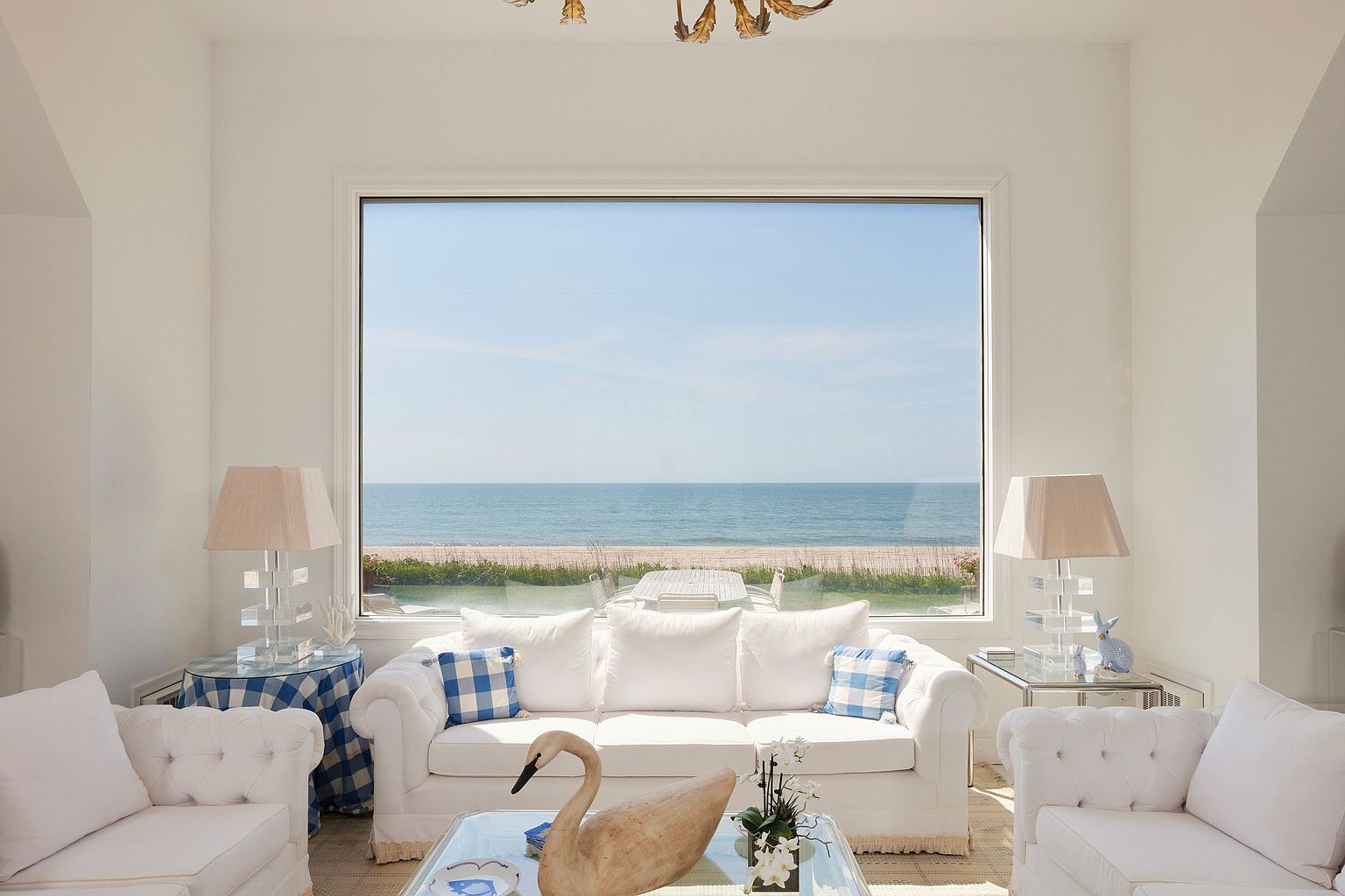 This is an interior shot of a bright and airy living room with a coastal aesthetic. The room features white sofas with tufted details and blue and white patterned throw pillows, complemented by glass-topped tables and a decorative swan sculpture. A large window provides a stunning view of the ocean and beach, enhancing the room's serene and luxurious atmosphere.