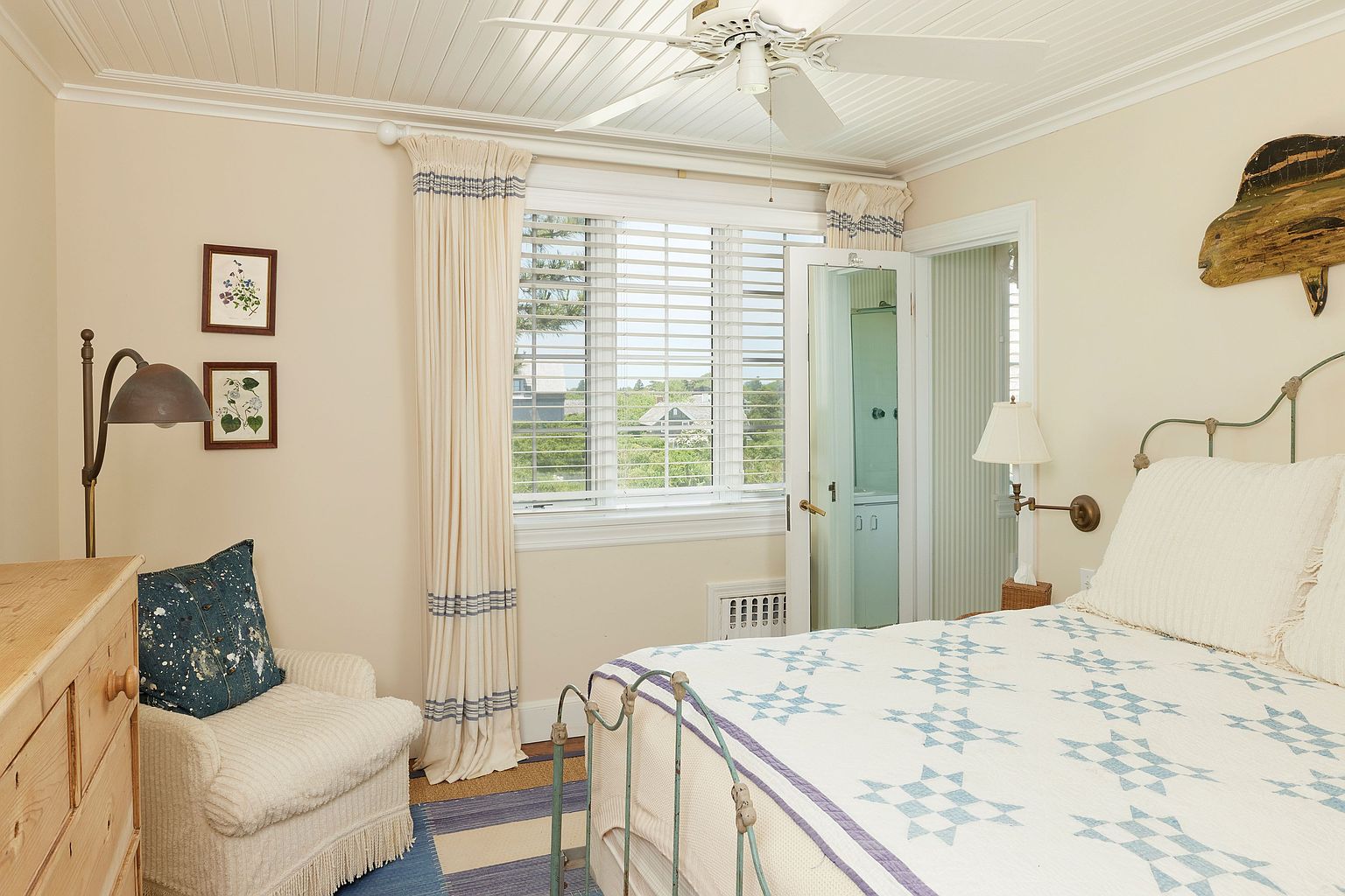This is a cozy guest bedroom featuring a bed with a blue and white quilt, a comfortable armchair, and a window with natural light. The room has a relaxed, cottage-style aesthetic with light-colored walls and a ceiling fan. The presence of artwork and a bedside lamp adds to the inviting atmosphere.