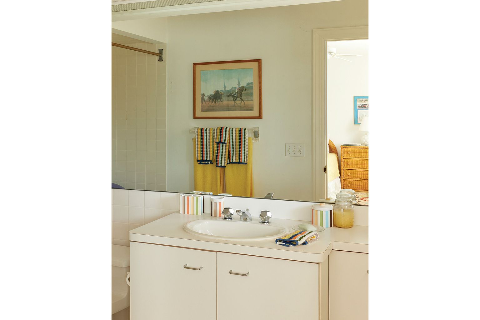 This bathroom features a white vanity with a built-in sink and chrome fixtures. A large mirror reflects the room, including a framed artwork above a towel rack with colorful towels. The overall impression is clean and well-maintained, with a touch of color.