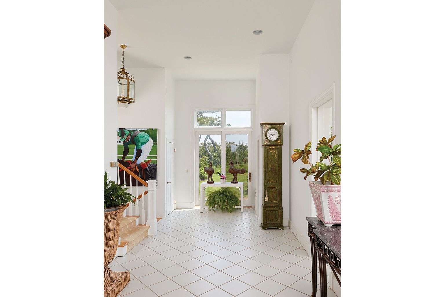 This is an interior shot of a bright and spacious hallway. The hallway features white walls, a white tile floor, and a staircase with white railings. A large painting hangs on the wall near the stairs, and a grandfather clock stands against the wall near a window, adding a touch of classic elegance to the space.