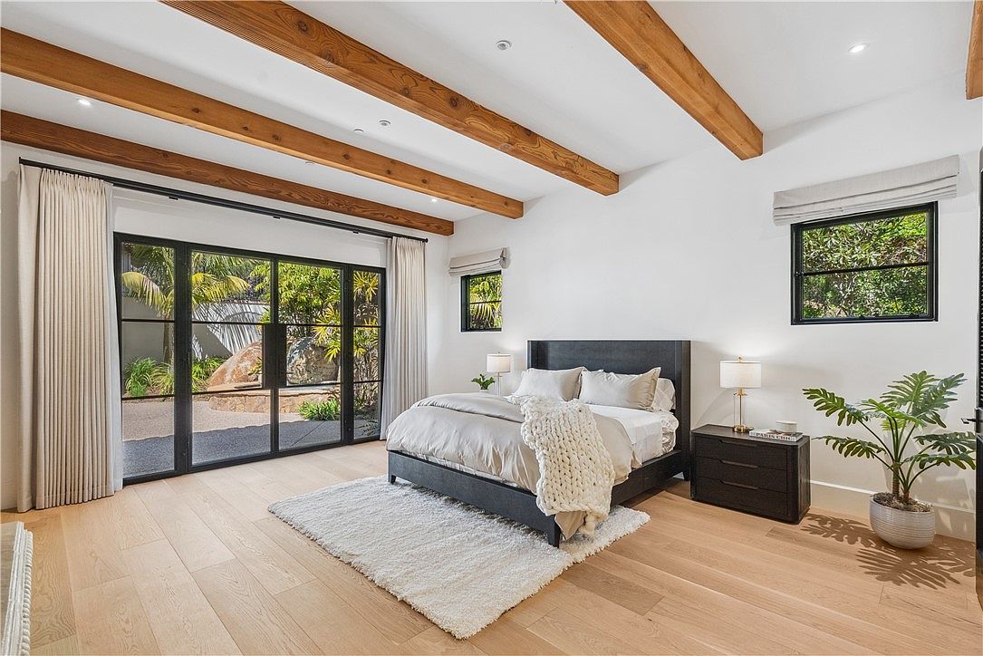 This is a bright and airy primary bedroom featuring a neutral color palette with wooden beam accents on the ceiling. The room is well-lit with natural light streaming through large windows and glass doors, offering a view of the outdoor greenery. A comfortable bed is centered in the room, complemented by a dark wood nightstand and a stylish area rug, creating a serene and inviting atmosphere.