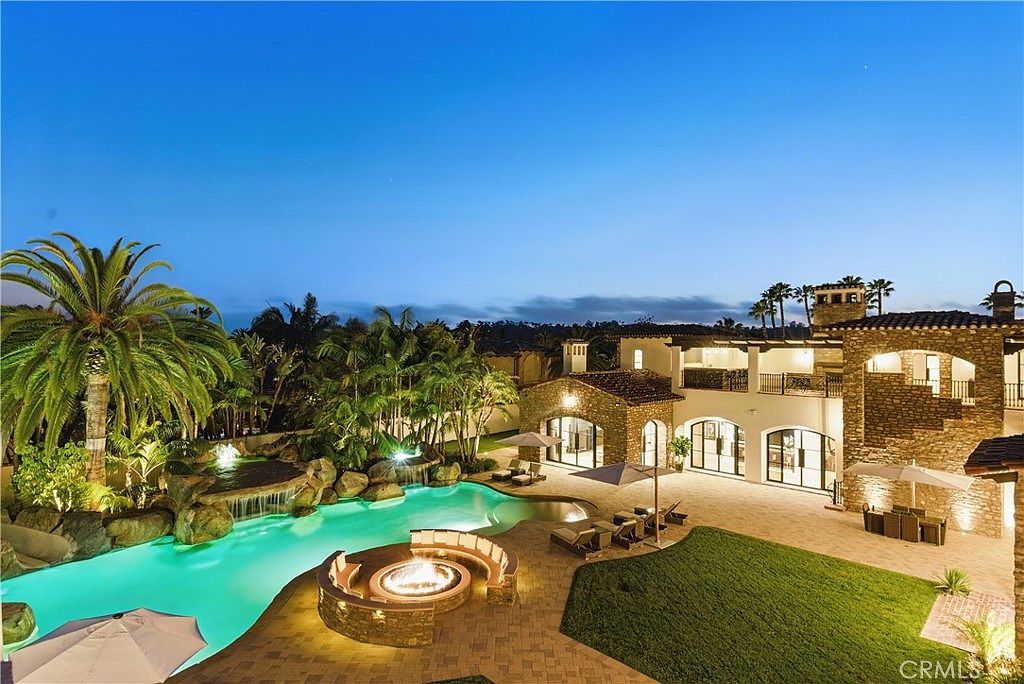 This is a stunning rear view of a luxurious estate at dusk, showcasing a resort-style backyard. The focal point is a large, lagoon-style pool with a waterfall feature, surrounded by lush landscaping and palm trees. A circular fire pit with built-in seating and a manicured lawn provide additional outdoor living spaces, complemented by the elegant architecture of the home with its stone accents and multiple balconies.