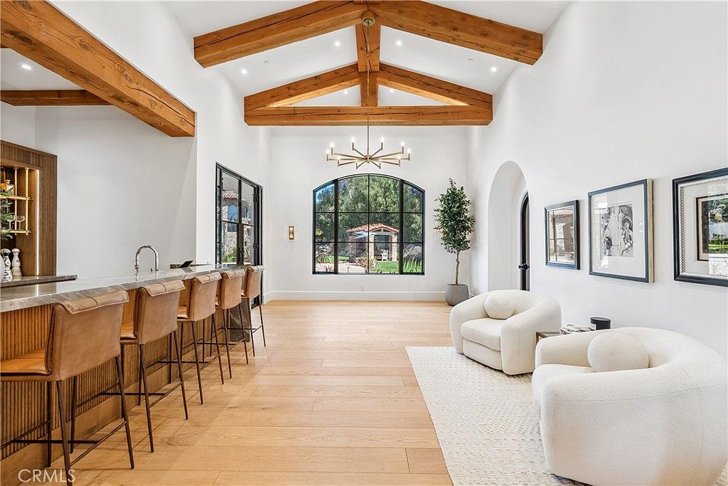 This is an interior shot of a living room featuring high ceilings with exposed wooden beams and a large arched window providing natural light. The room is furnished with two modern, rounded armchairs and a long bar with stools, creating a blend of living and entertaining spaces. The overall impression is bright, spacious, and stylish.