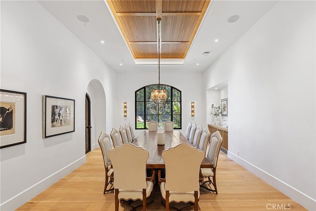 This is a dining room featuring a long wooden table with upholstered chairs, a decorative chandelier, and a large arched window. The walls are painted white, and the floors are hardwood. The room has a formal and elegant style, suitable for large gatherings.