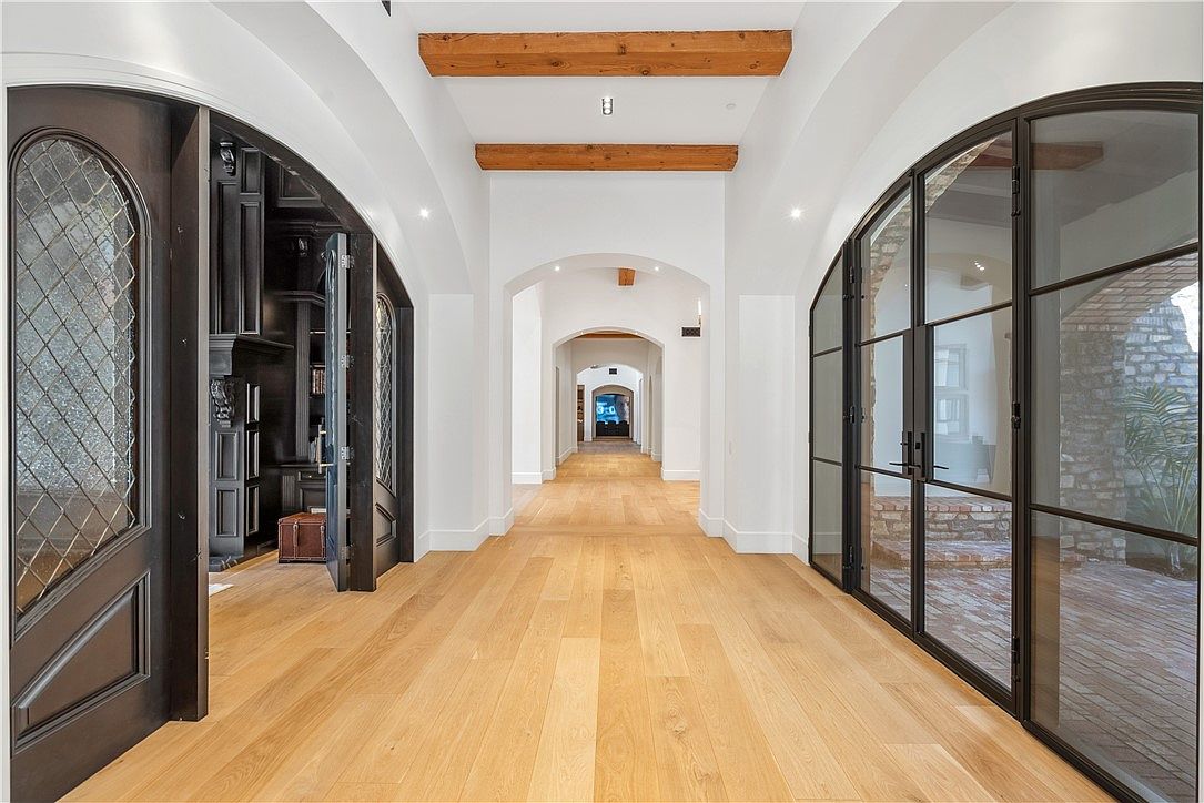 This is an interior shot of a long hallway featuring light hardwood flooring and white walls. The hallway is punctuated by arched doorways and wooden beams on the ceiling, creating a sense of depth and architectural interest. On either side, there are dark wood doors and large windows with black frames, adding contrast and visual appeal.