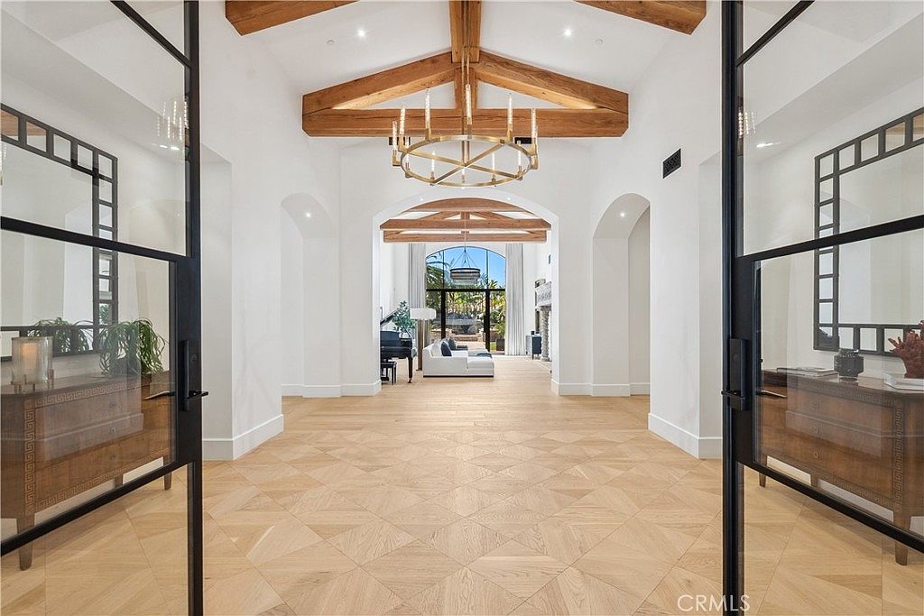 This grand hallway features a stunning parquet wood floor, white walls, and arched doorways leading to other rooms. Exposed wooden beams and a circular chandelier add architectural interest. The open doors at the entrance provide a glimpse into the home's interior, creating a welcoming and luxurious atmosphere.