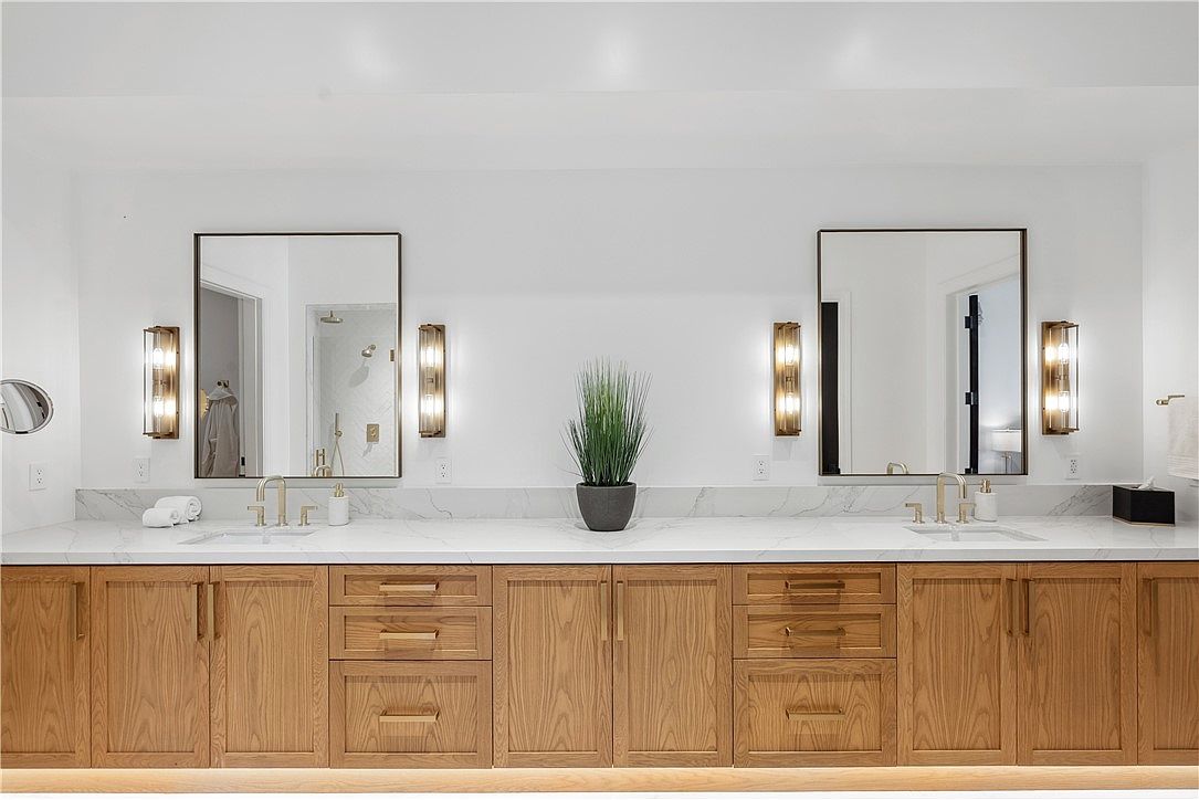 This is a well-lit primary bathroom featuring a double vanity with light wood cabinetry and a white marble countertop. Two rectangular mirrors are mounted above the sinks, flanked by modern sconces. A potted plant sits in the center of the vanity, adding a touch of nature to the space.
