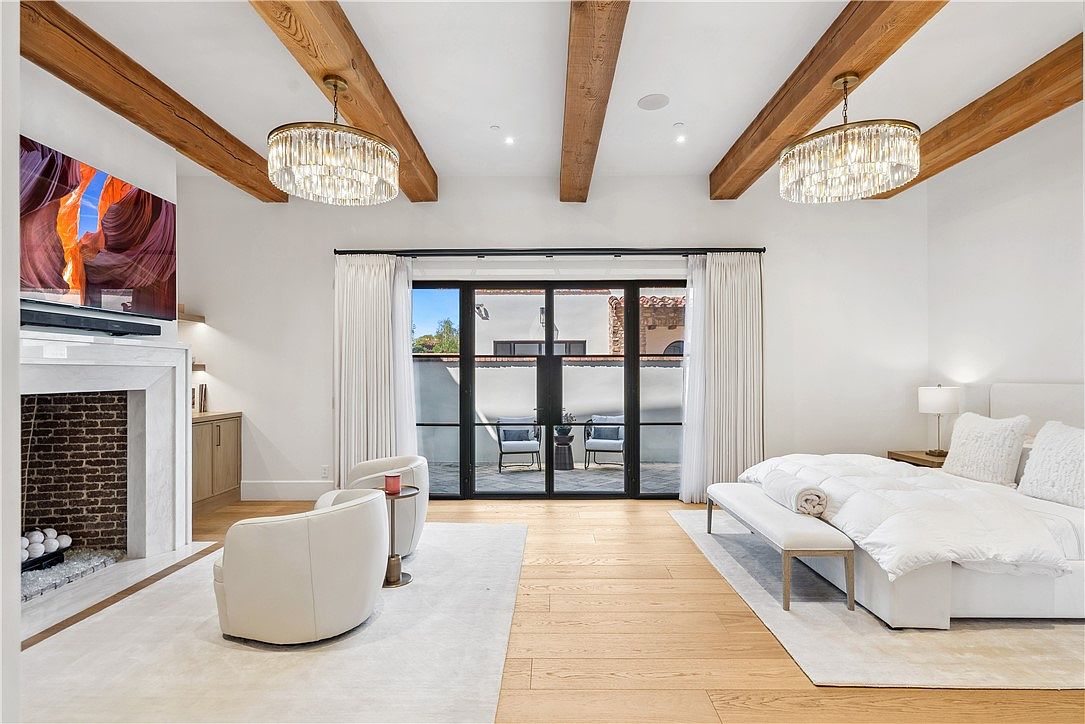 This is a bright and airy primary bedroom featuring exposed wooden beams, a modern fireplace with a TV above, and a luxurious bed with white linens. French doors lead to an outdoor patio, enhancing the indoor-outdoor flow. The room is decorated in a neutral color palette, creating a serene and inviting atmosphere.