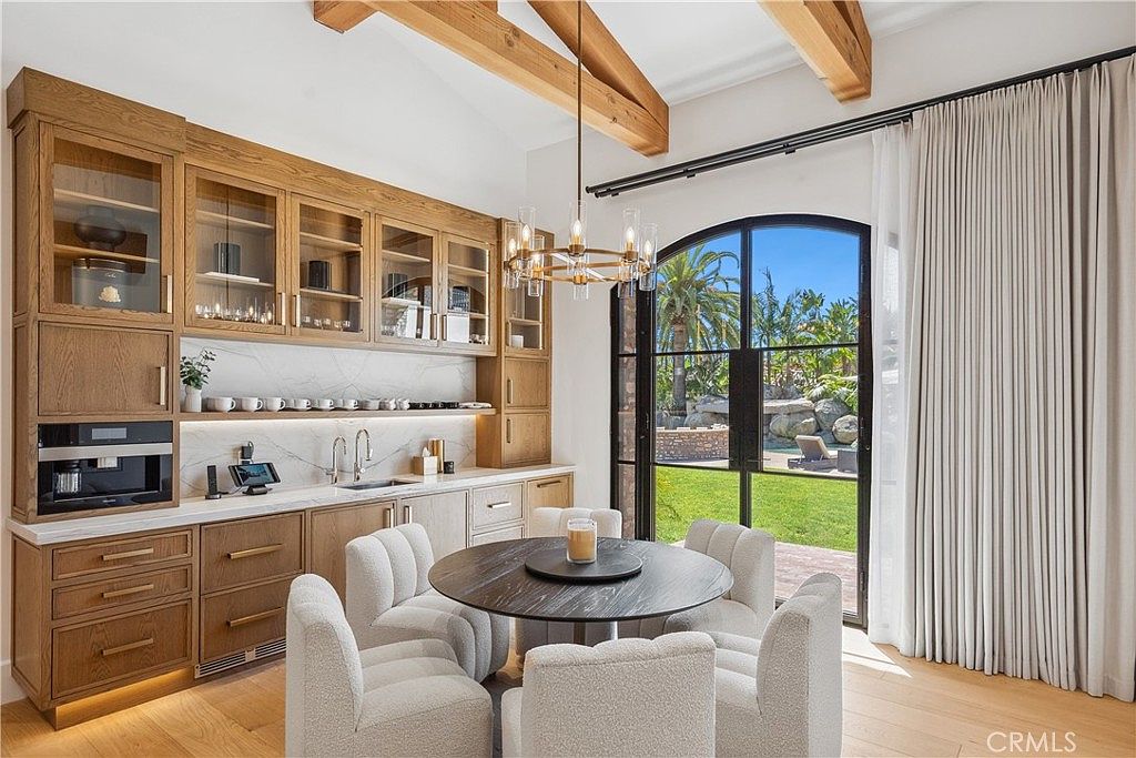This interior shot showcases a stylish dining area featuring a round dark wood table surrounded by six white upholstered chairs. Built-in wooden cabinetry with glass doors lines one wall, complemented by marble countertops and backsplash. A modern chandelier hangs above the table, and a large arched window with black frames offers a view of the outdoor landscape, enhancing the room's bright and airy atmosphere.